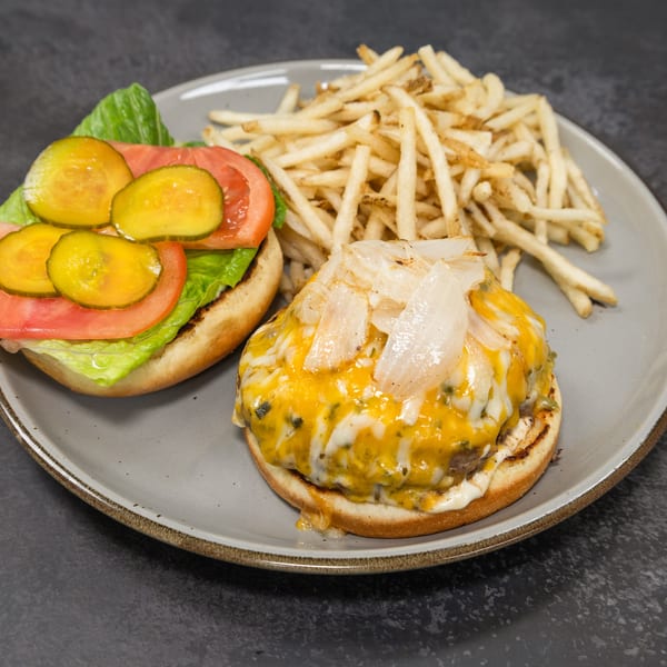 Open burger with melted cheese, caramelized onions, lettuce, tomato, pickles, and french fries on a gray plate.