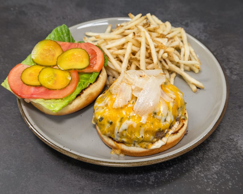 Open burger with melted cheese, caramelized onions, lettuce, tomato, pickles, and french fries on a gray plate.