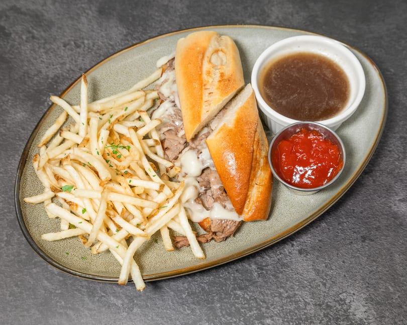 Philly cheesesteak sandwich with fries, au jus, and ketchup on a speckled plate.