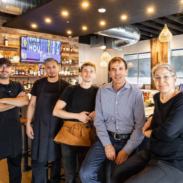 Five smiling restaurant staff members, some in aprons, pose behind a well-stocked bar.