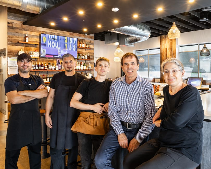 Five smiling restaurant staff members, some in aprons, pose behind a well-stocked bar.