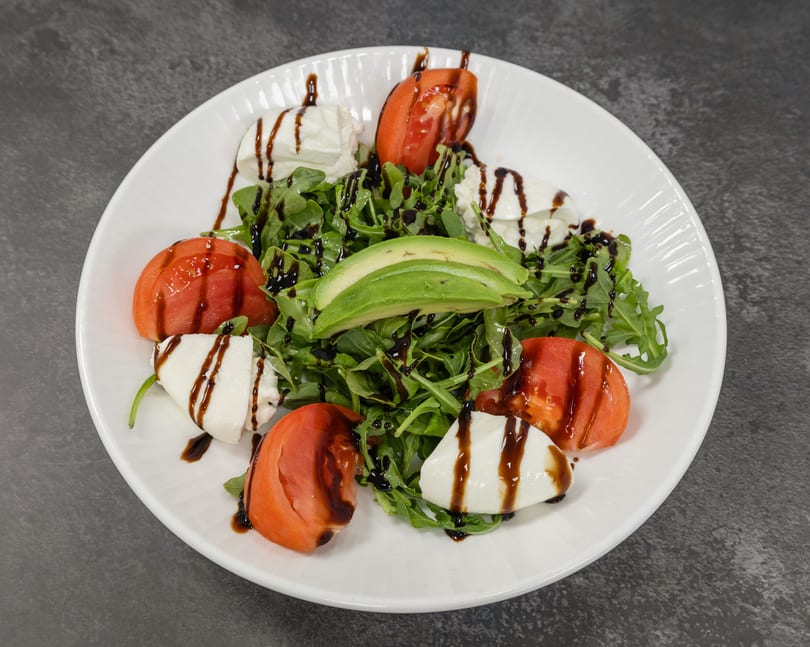 Fresh arugula salad with tomatoes, mozzarella, avocado, and balsamic drizzle on a white plate.