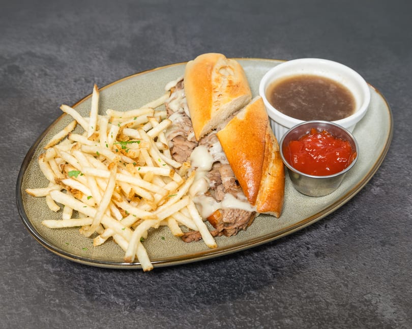 Cheesy French dip sandwich with golden fries, au jus, and ketchup on a grey plate.