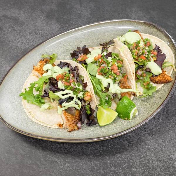 Three vibrant fish tacos topped with fresh salsa, green sauce, and lime on a grey plate.