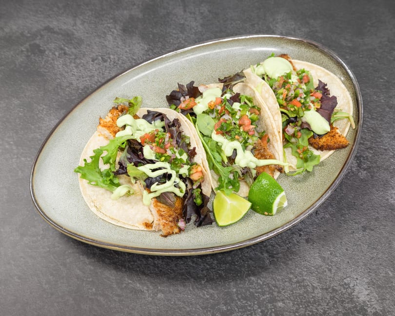 Three vibrant fish tacos topped with fresh salsa, green sauce, and lime on a grey plate.