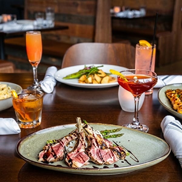 A restaurant table set with various dishes including lamb chops, pasta, skewers, and cocktails.