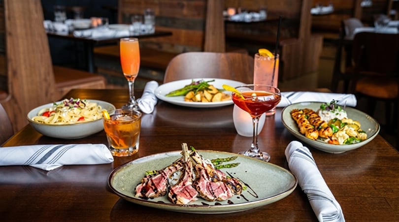 A restaurant table set with various dishes including lamb chops, pasta, skewers, and cocktails.