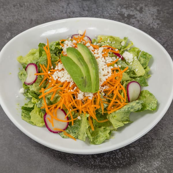 A vibrant green salad with avocado, shredded carrots, crumbled cheese, and radish slices in a white bowl.
