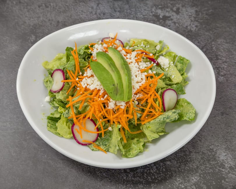 A vibrant green salad with avocado, shredded carrots, crumbled cheese, and radish slices in a white bowl.