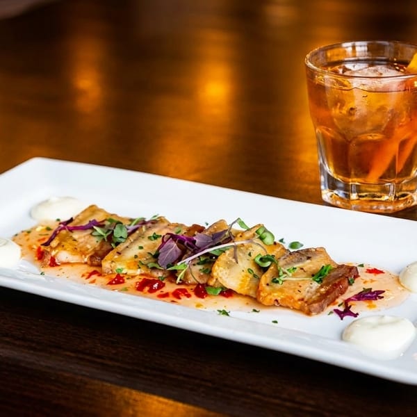 Appetizer of seared scallops with sauce and herbs, alongside a cocktail, on a dark wooden table.
