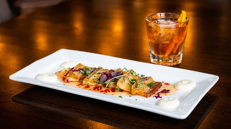 Appetizer of seared scallops with sauce and herbs, alongside a cocktail, on a dark wooden table.