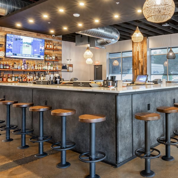 Modern restaurant bar with a long counter, stools, liquor shelves, a TV, and woven pendant lights.