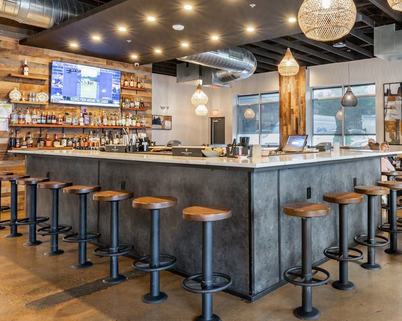 Modern restaurant bar with a long counter, stools, liquor shelves, a TV, and woven pendant lights.