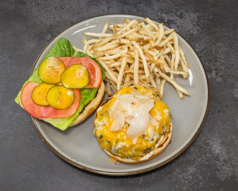 Open-faced cheeseburger with grilled onions, lettuce, tomato, pickles, and a side of crispy french fries.