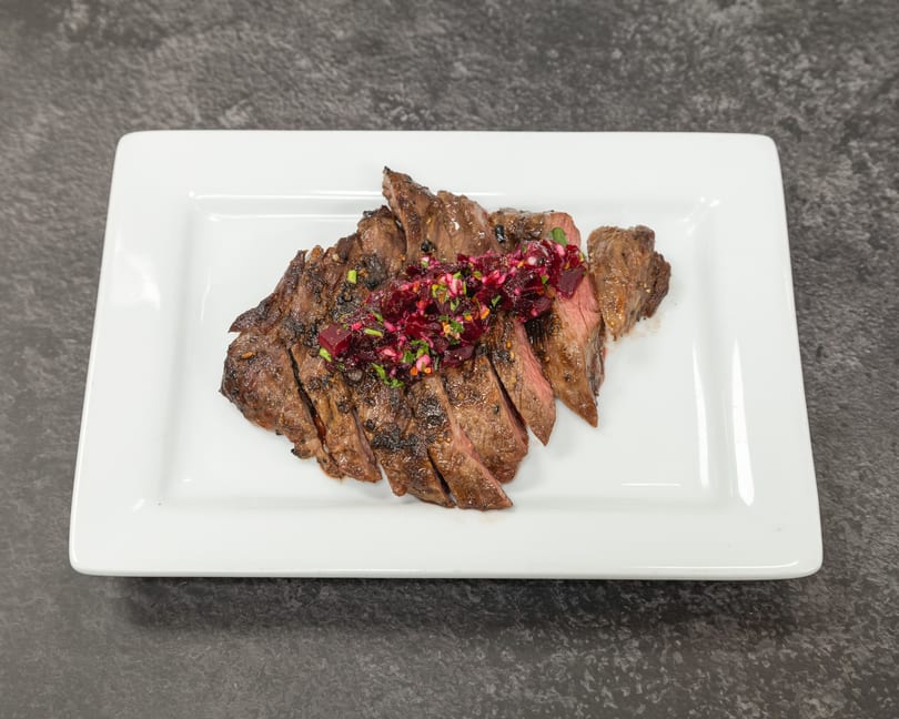 Sliced grilled steak topped with a vibrant red beet relish and green herbs on a white rectangular plate.